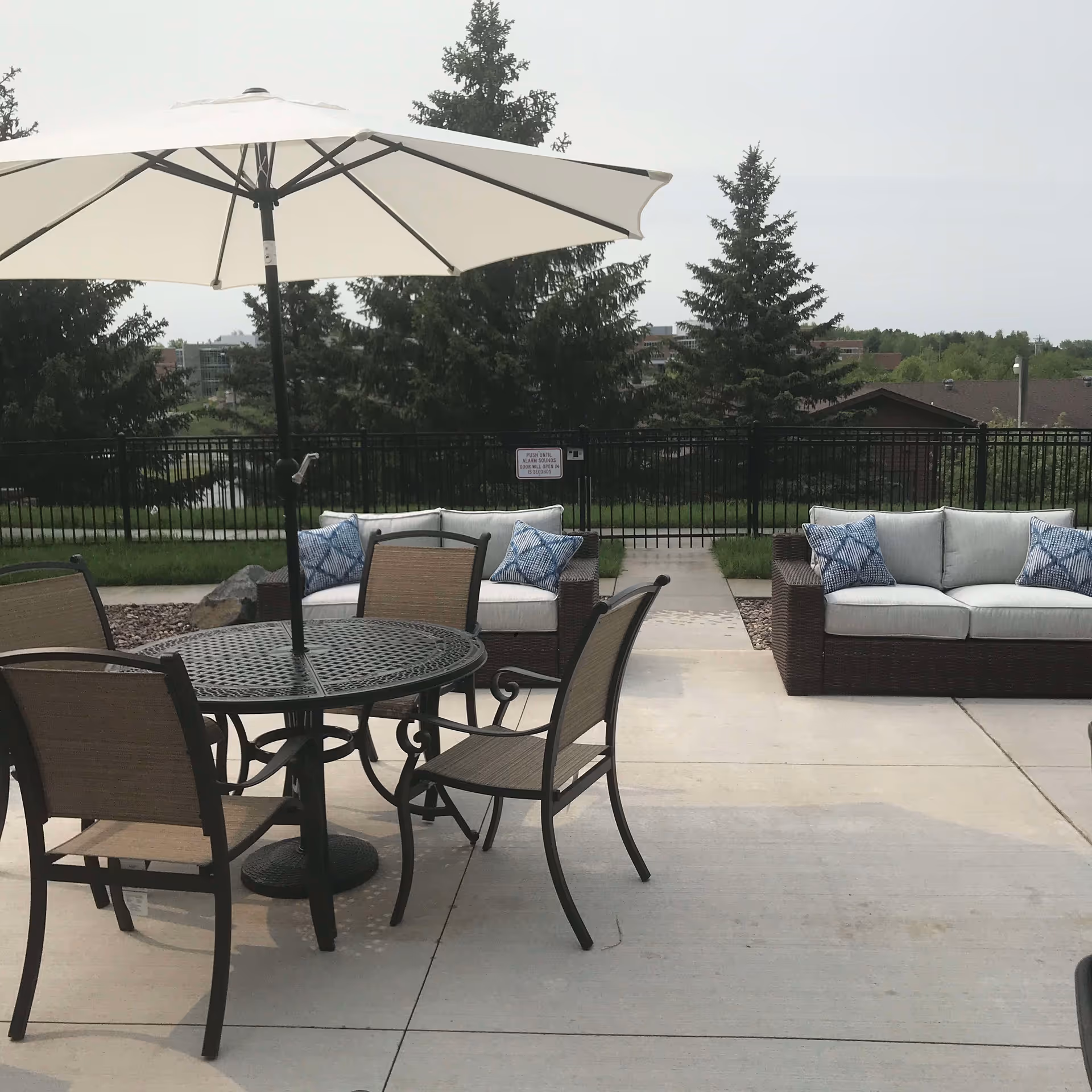 Outdoor patio with a round metal table and umbrella, several chairs, and wicker sofas with cushions in front of a fenced lawn and trees.