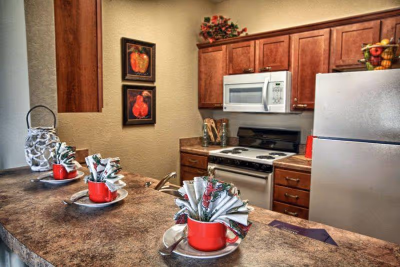 Interior view of a kitchen with wooden cabinets, a white microwave above a stove, and a refrigerator. A countertop in the foreground has three red cups with decorative folded napkins placed on white plates. Two framed pictures of fruit hang on the wall.