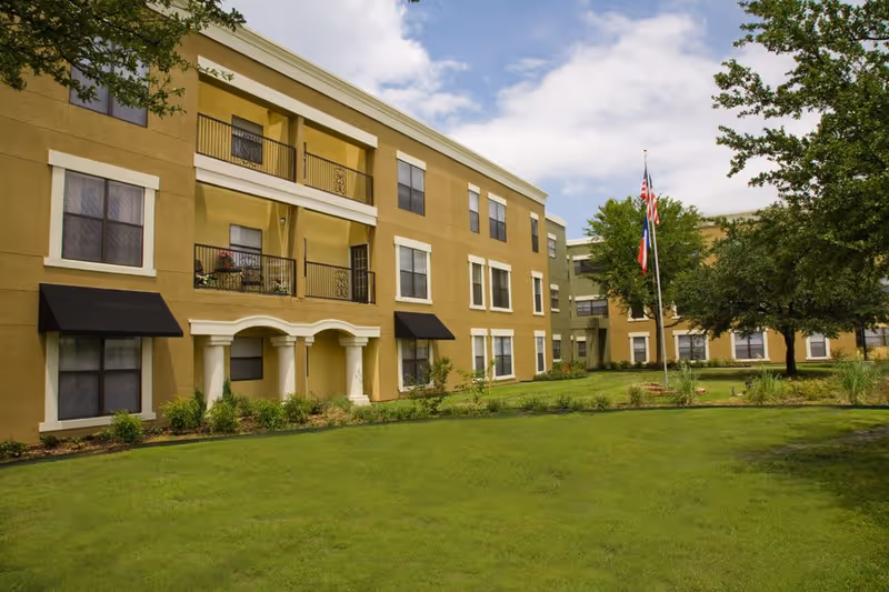 Exterior view of a three-story assisted living facility building with beige and green walls, black window awnings, balconies, and a well-maintained lawn with trees and flagpoles displaying the American and Texas flags under a partly cloudy sky.