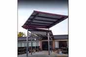 Covered entrance area with a metal roof structure supported by beams in front of a building with large windows and a flat roof, surrounded by some plants and outdoor seating.