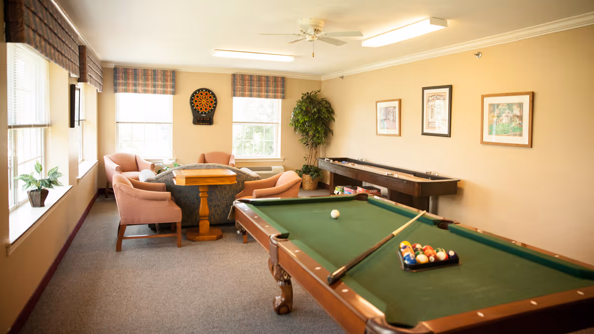 A bright communal game room with a pool table, seating area, dartboard, and shuffleboard table.