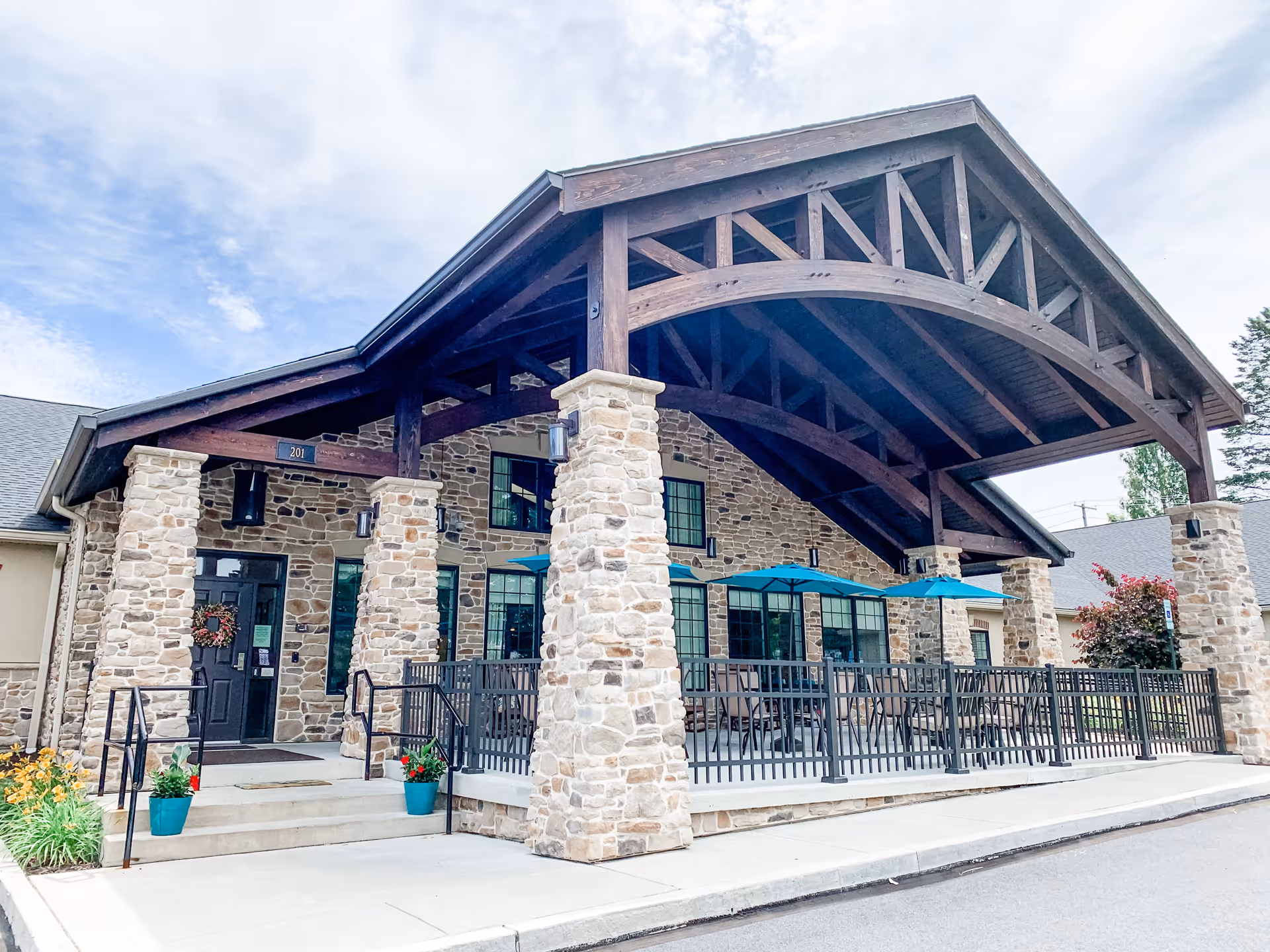 Stone-clad building front with a large timbered porte-cochère, entrance steps and an outdoor patio with blue umbrellas.