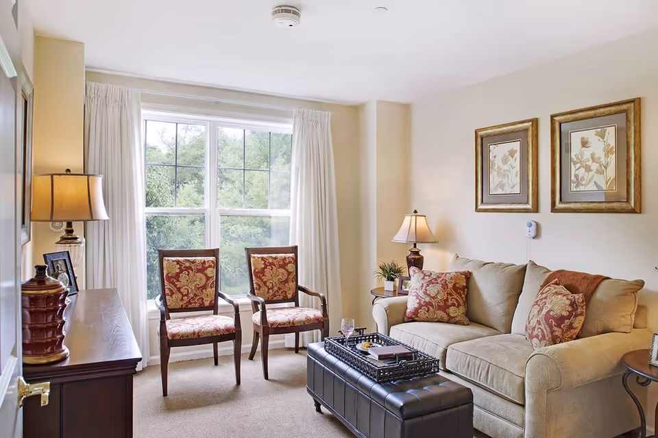 A cozy living room with a beige sofa adorned with patterned cushions, two wooden chairs with red and gold upholstery, a dark leather ottoman serving as a coffee table with a tray on top, two table lamps providing warm lighting, framed floral artwork on the wall, and a large window with white curtains letting in natural light and showing greenery outside.