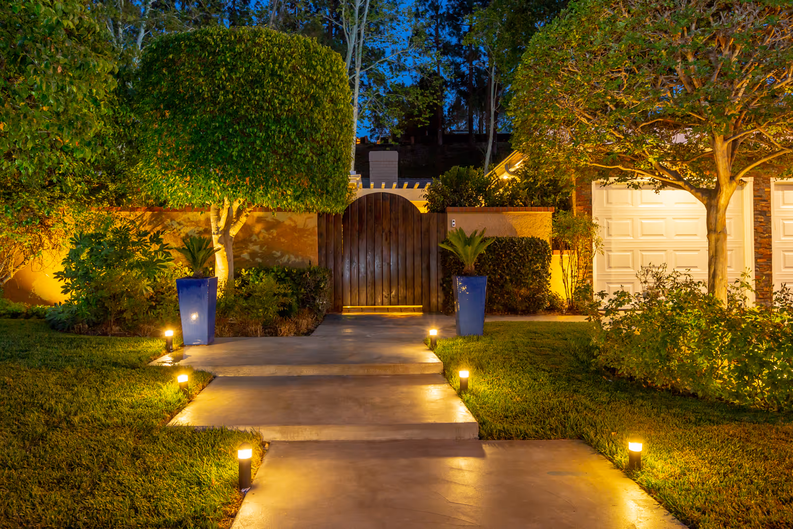 A well-lit outdoor pathway at dusk leading to a wooden gate surrounded by lush greenery and two large blue planters with small palm plants. The pathway is flanked by small ground lights and bordered by neatly trimmed grass and trees. A white garage door is visible on the right side.