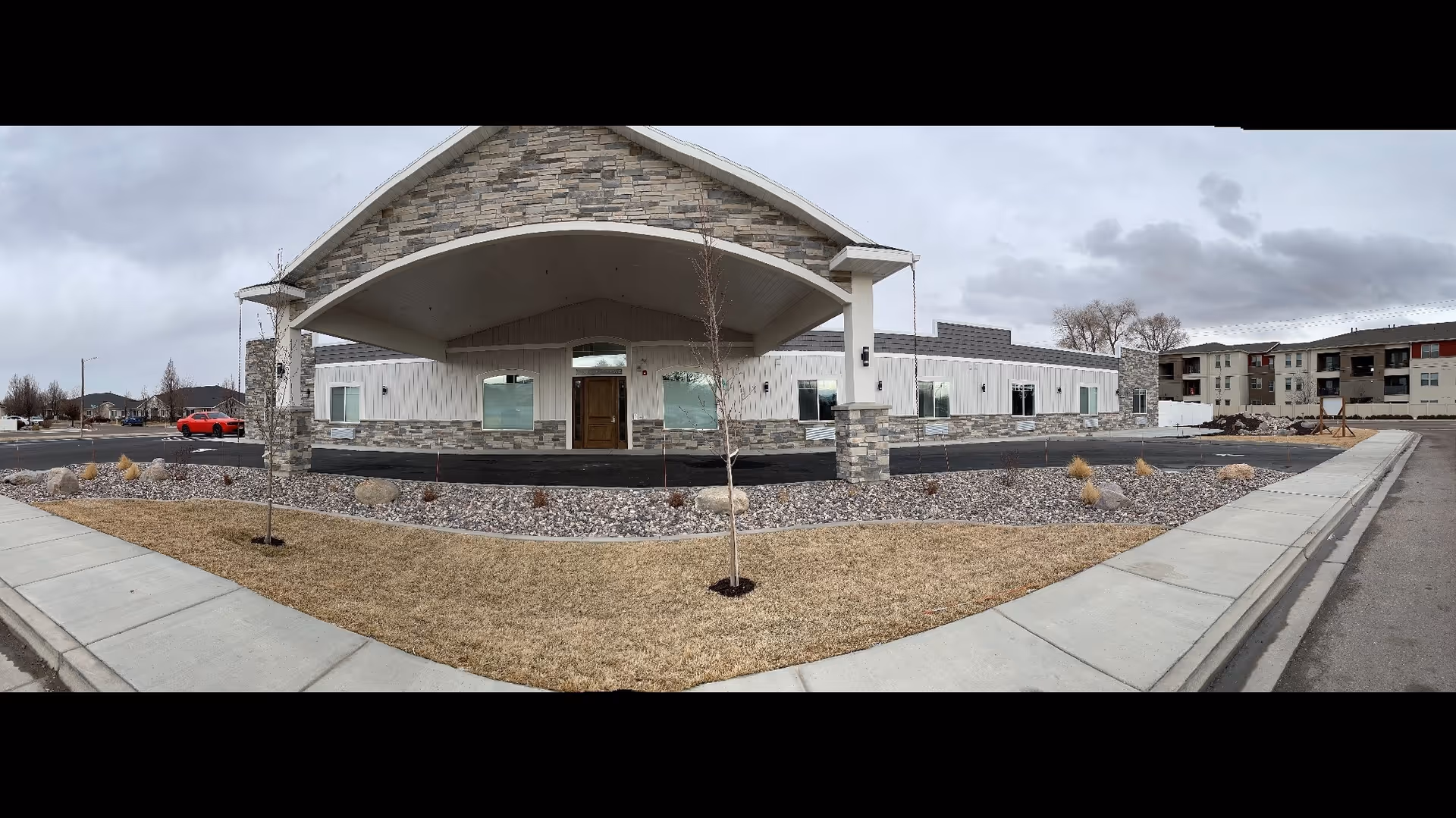 Front exterior view of a single-story residential building with stone and white siding, a covered entrance with a wooden door, small trees and landscaping with rocks and grass, and a sidewalk in front. Overcast sky in the background.