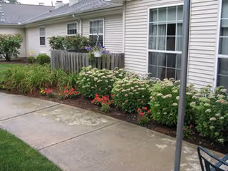 Outdoor view of a garden area next to a building with beige siding and multiple windows. The garden features various flowering plants and shrubs along a concrete walkway.