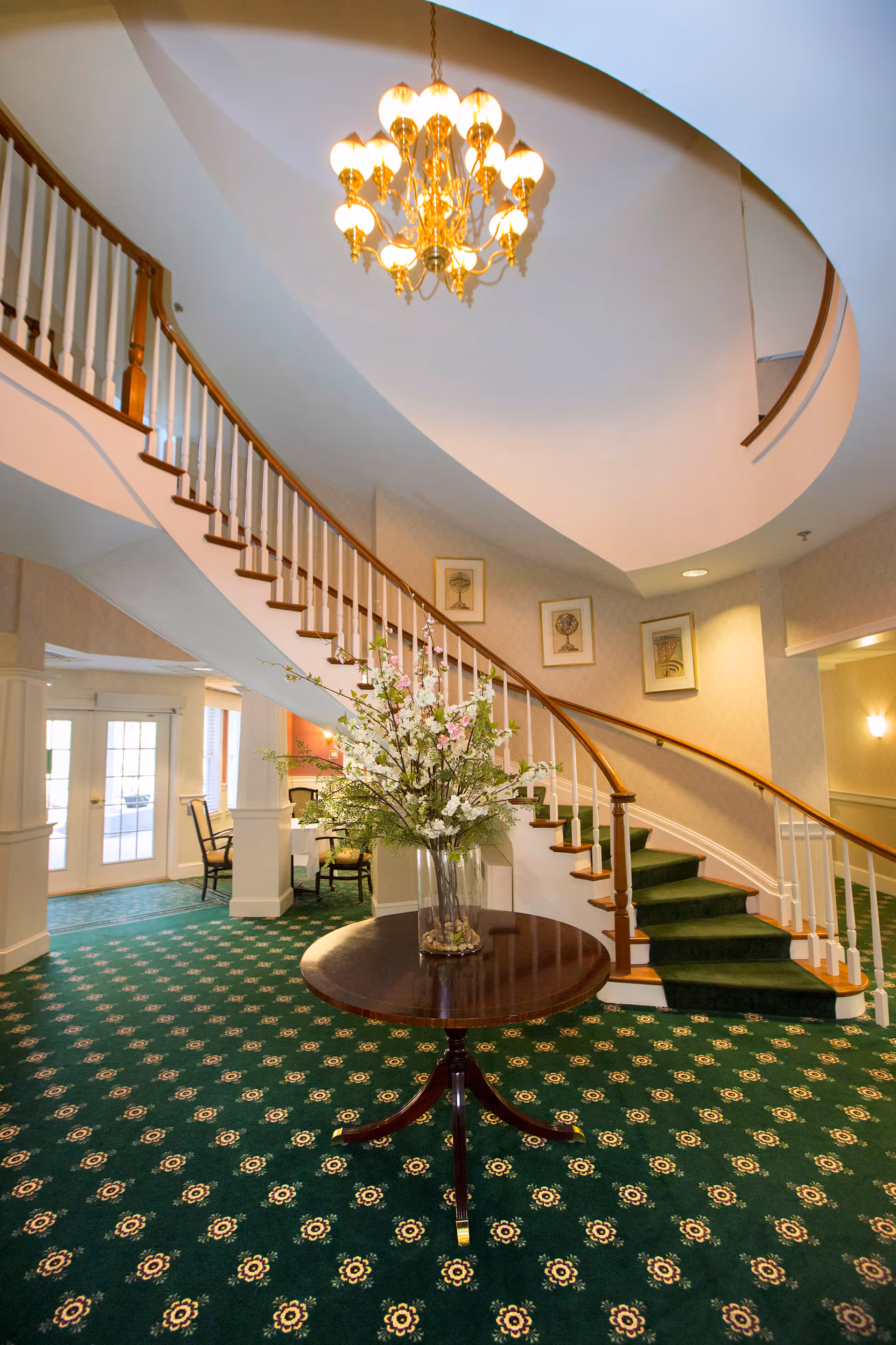 Interior view of a senior living facility featuring a curved staircase with wooden handrails and green carpeted steps. A round wooden table with a large floral arrangement is centered in front of the staircase. The floor is covered with green patterned carpet, and framed artwork is displayed on the wall along the staircase. A chandelier with multiple lights hangs from the ceiling, and a seating area with chairs is visible in the background near glass doors.
