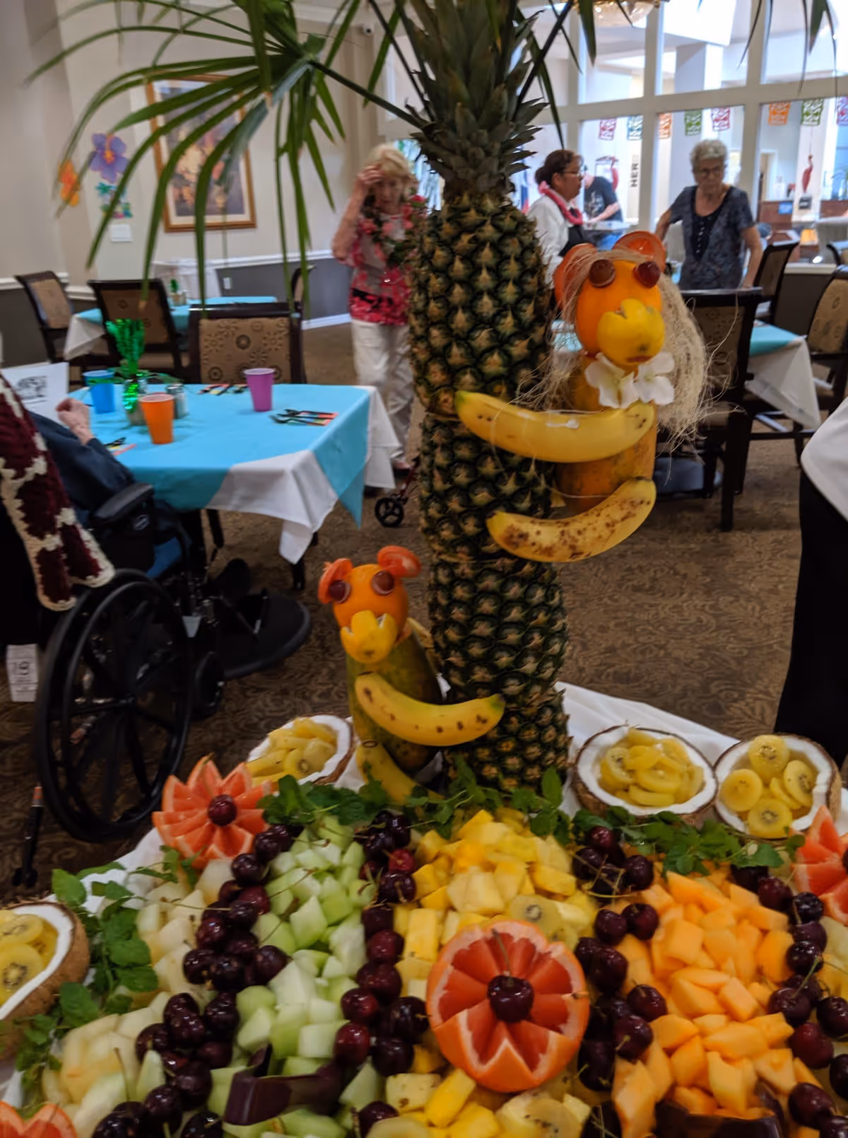 A colorful fruit display in a dining area featuring creatively arranged fruits including pineapples, bananas, cherries, melon, and kiwi. The centerpiece includes two animal figures made from fruits hugging a pineapple. In the background, elderly people are seen standing and sitting around tables covered with blue tablecloths.