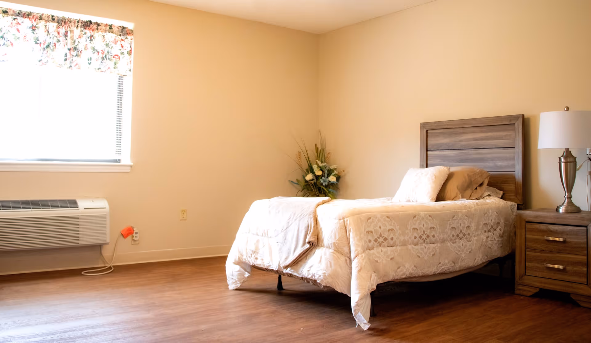 A simple bedroom with a single bed covered in a beige patterned comforter and pillows. Next to the bed is a wooden nightstand with a silver lamp. There is a window with floral curtains and an air conditioning unit below it. A floral arrangement is placed in the corner of the room.