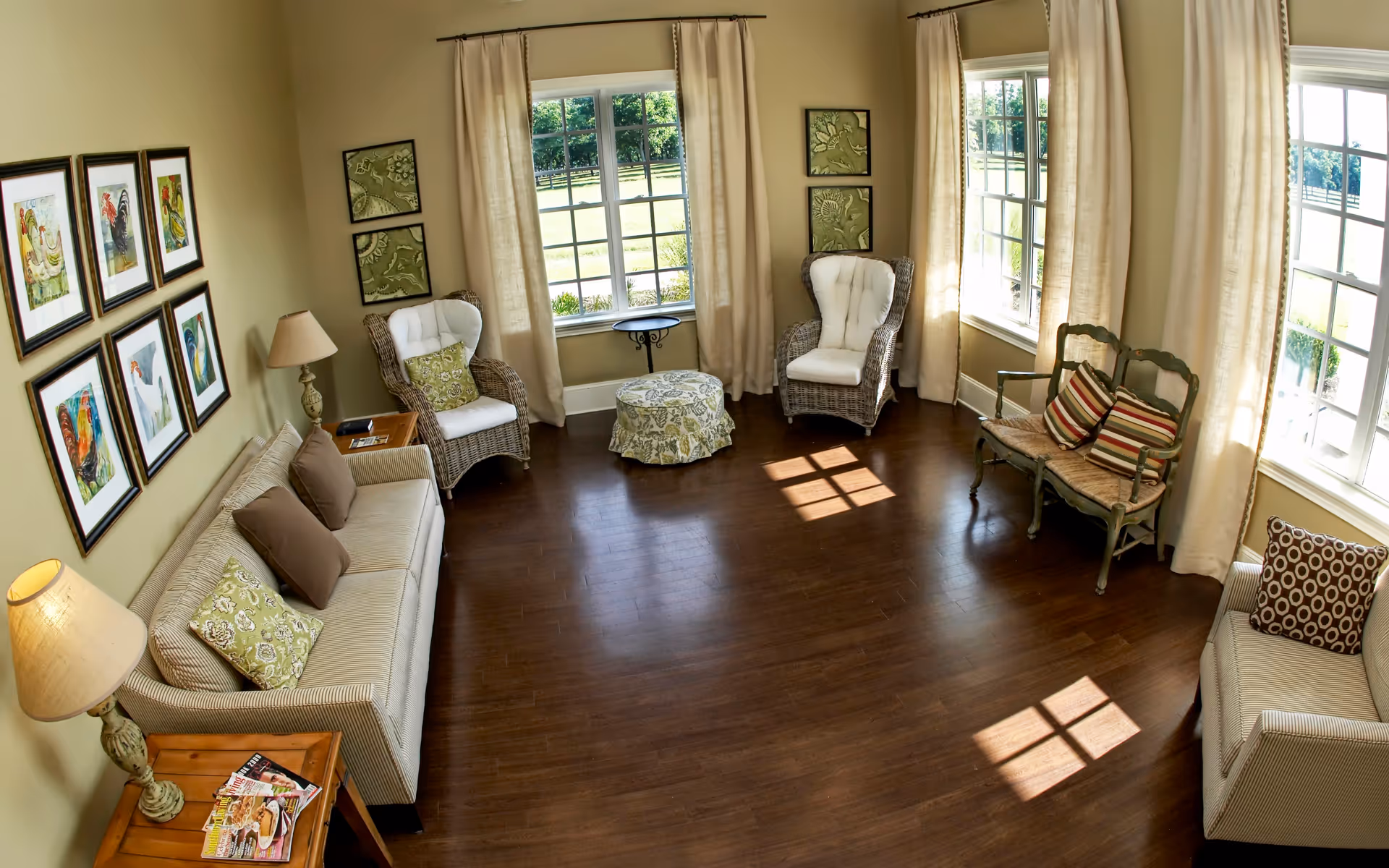 A bright and cozy living room with large windows letting in natural light. The room features two beige sofas with decorative pillows, two wicker armchairs with cushions, a small round ottoman, a wooden side table with a lamp and magazines, and framed artwork on the walls. Light-colored curtains frame the windows, and the floor is dark hardwood.