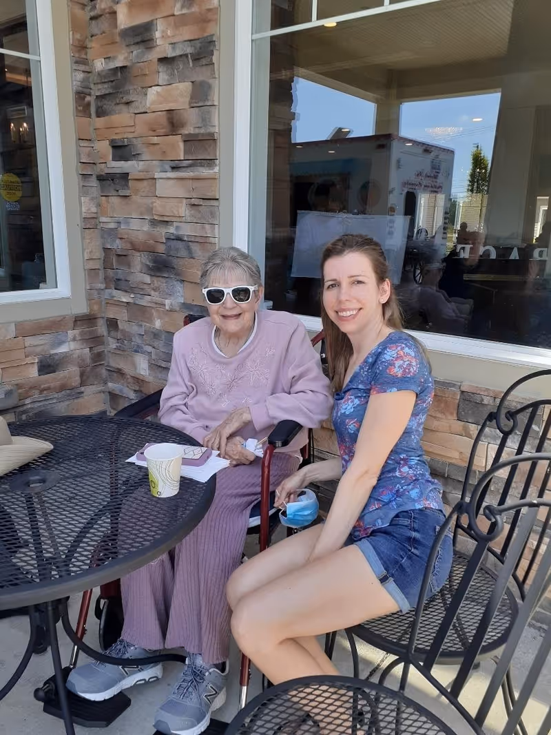 An elderly woman wearing sunglasses and a light purple outfit sits in a wheelchair next to a younger woman in a blue floral shirt and denim shorts. They are seated outside at a black metal table and chairs on a patio with a stone wall and windows behind them. The elderly woman has a paper cup and some papers on the table in front of her.