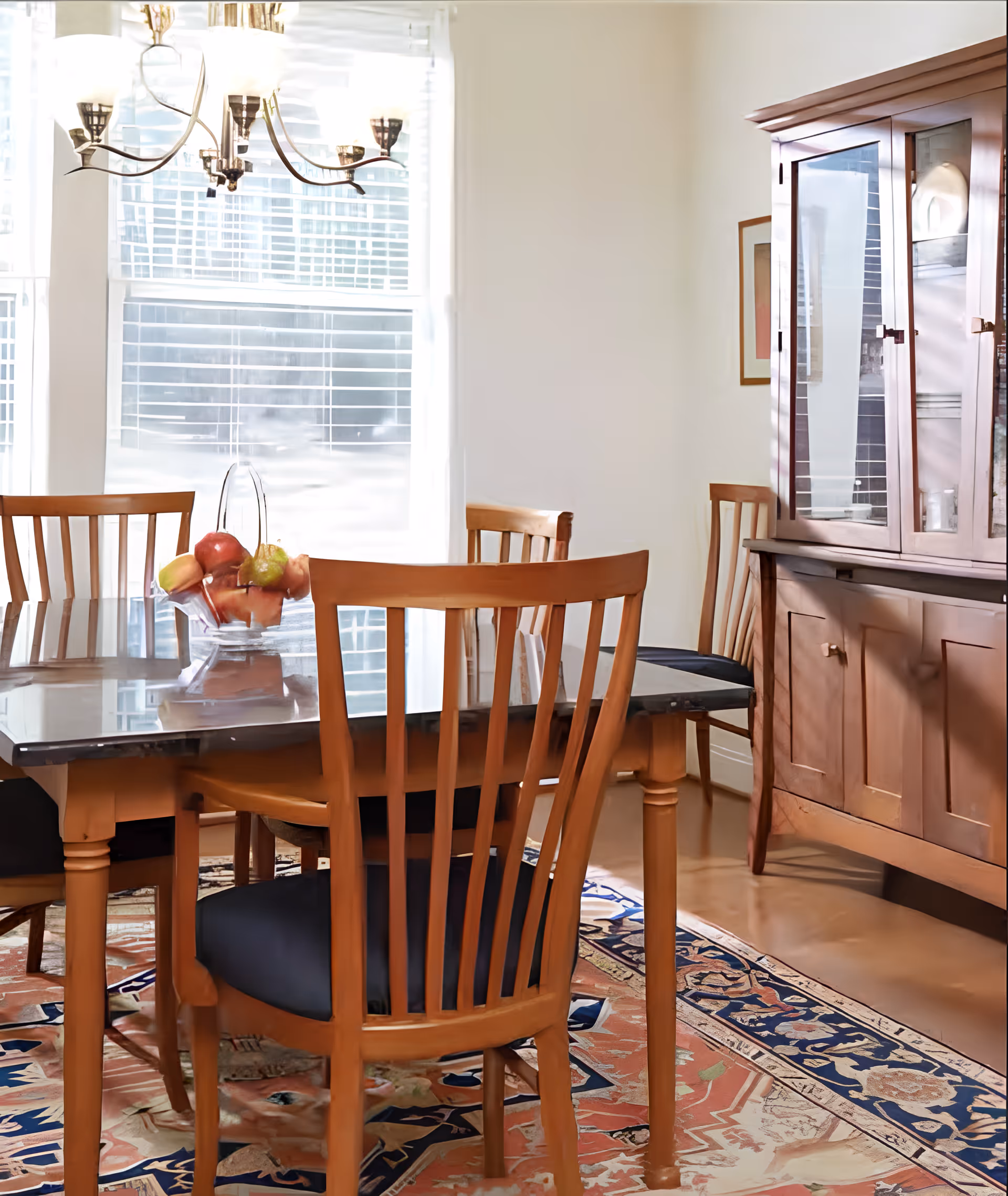 A bright dining room with a wooden table and matching chairs with dark cushions. A glass bowl filled with fruit sits on the table. There is a wooden china cabinet against the wall and a large window with white blinds letting in natural light. A decorative rug with intricate patterns covers the floor.