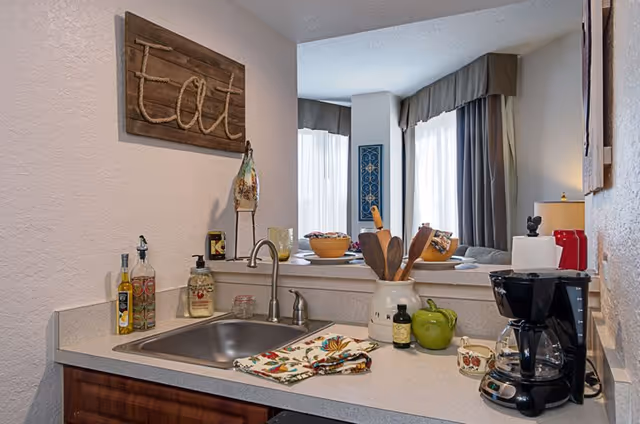 A small kitchen area with a stainless steel sink, countertop with various items including a coffee maker, decorative jars, a green apple-shaped container, and wooden utensils in a ceramic holder. A wooden wall decoration with the word 'Eat' hangs above the sink. In the background, there is a dining area with a table set with bowls and placemats, and large windows with curtains letting in natural light.