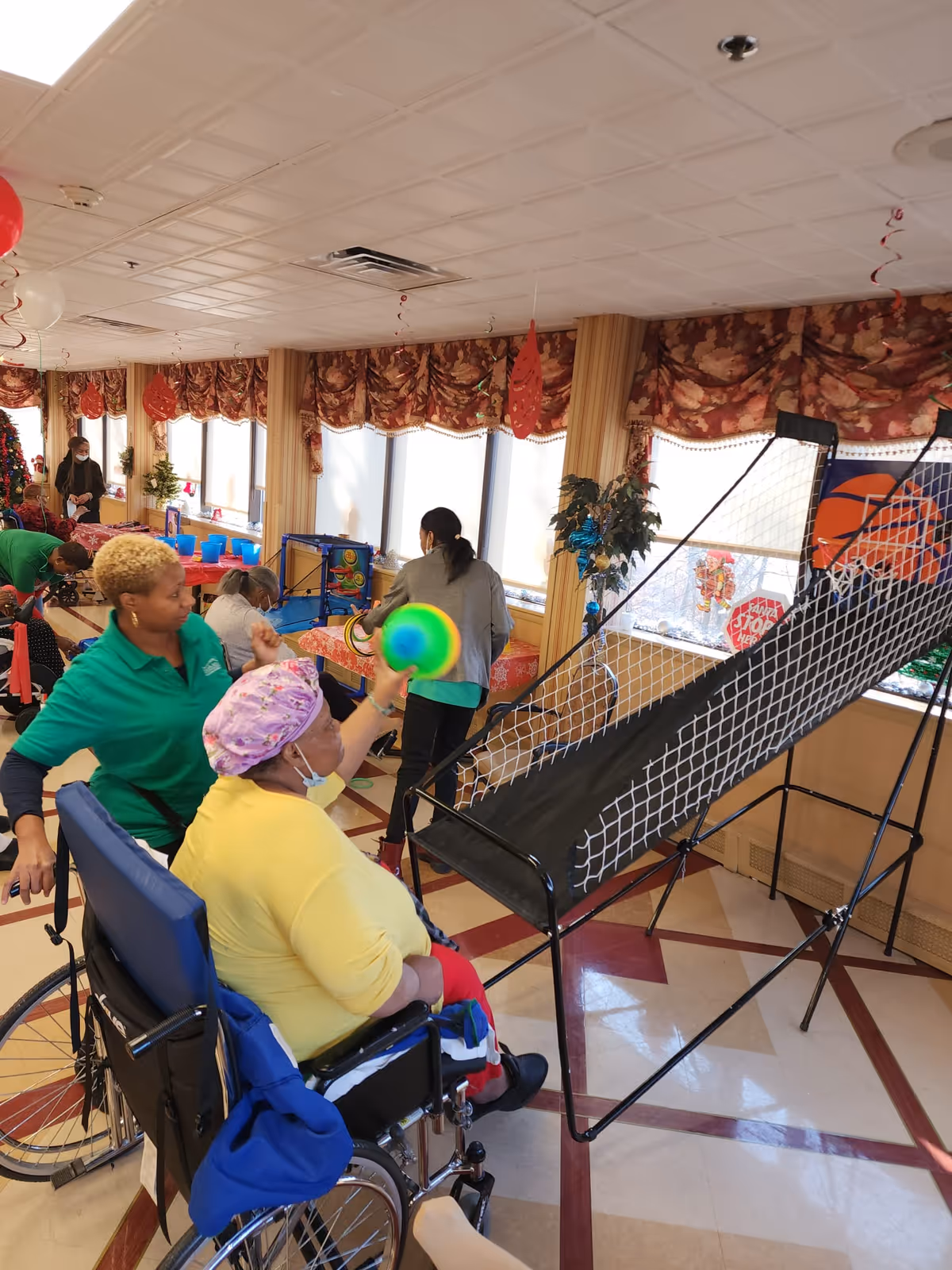 A group of people in a decorated room with large windows and patterned curtains. One person in a wheelchair wearing a yellow shirt and purple headscarf is playing a basketball arcade game, throwing a colorful ball towards the hoop. Another person in a green shirt is standing beside the wheelchair, assisting. Other people are visible in the background, engaging in various activities.