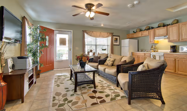 A cozy living room and kitchen area in a senior living facility with a beige sofa and two matching armchairs around a glass coffee table on a patterned rug. The kitchen has wooden cabinets, a white refrigerator, stove, microwave, and coffee maker. A ceiling fan with lights is above, and a red front door is open to a small porch area. A flat-screen TV is mounted on the wall opposite the seating area.