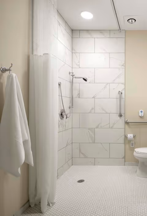 A clean, accessible bathroom with a walk-in shower featuring white marble tiles on the walls and small hexagonal white tiles on the floor. The shower has a handheld showerhead and a grab bar. A white shower curtain hangs on the left side, and a white towel is hanging on a hook on the wall. There is a toilet with a grab bar and a toilet paper holder on the right side.