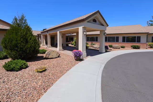 Exterior view of Park Manor Rehabilitation Center showing a covered entrance with columns, a curved driveway, and landscaped areas with rocks, bushes, and flowers under a clear blue sky.