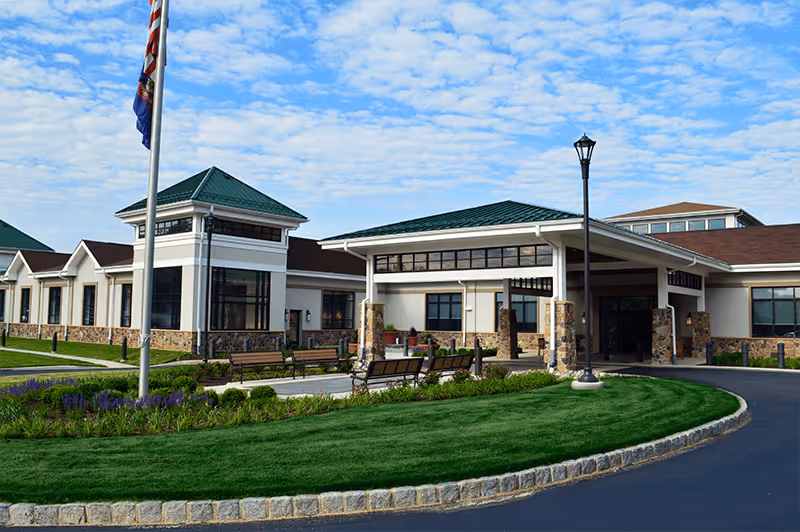 Exterior view of Granite Farms Estates senior living facility showing a single-story building with a green roof, stone and white walls, a covered entrance, flagpole, benches, landscaped green lawn, and a clear blue sky with scattered clouds.