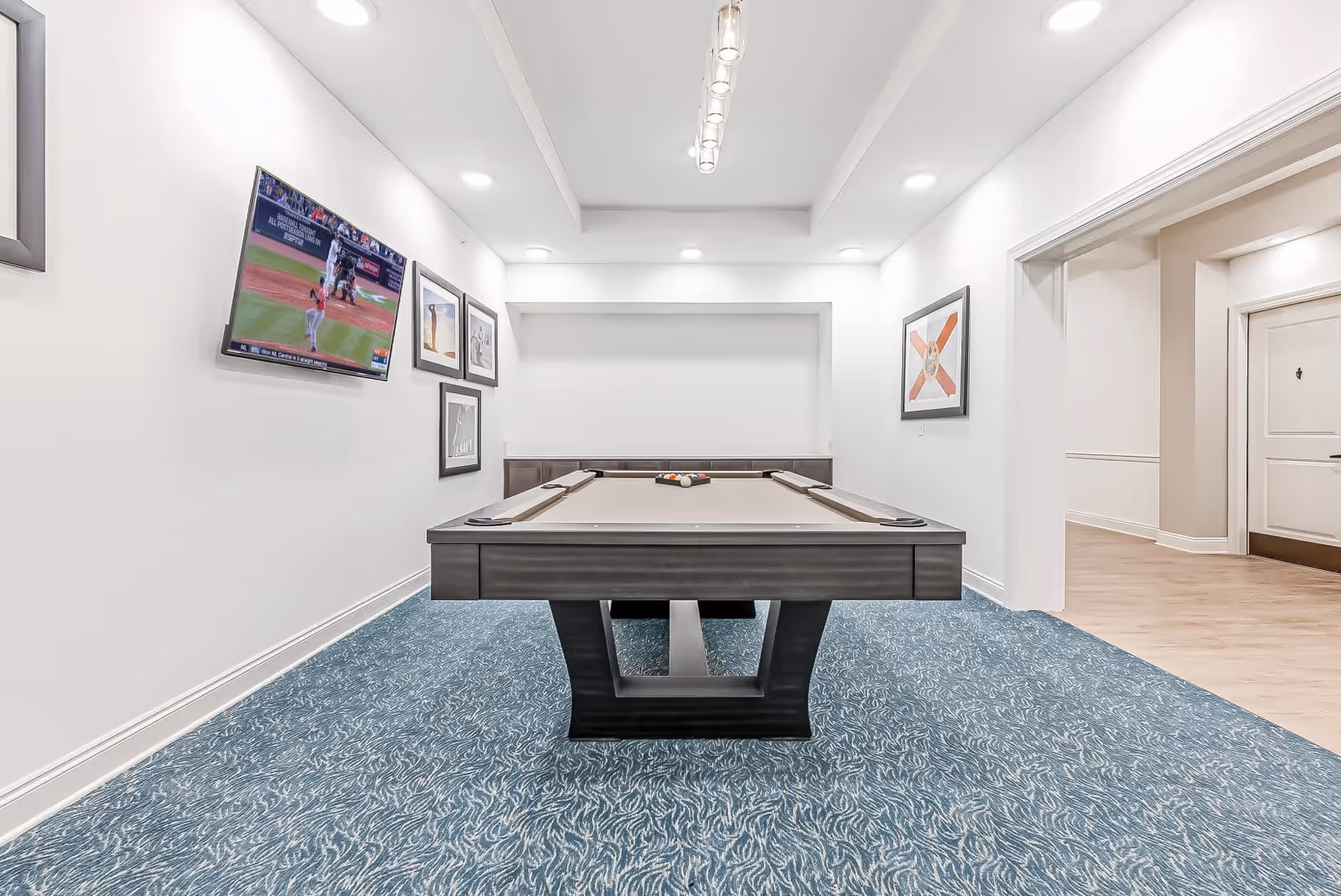 A modern game room featuring a pool table with balls arranged for a game. The room has white walls adorned with framed pictures and a flat-screen TV mounted on the left wall showing a baseball game. The floor is covered with a blue patterned carpet, and recessed lighting illuminates the space. An open doorway leads to a hallway with light wood flooring.