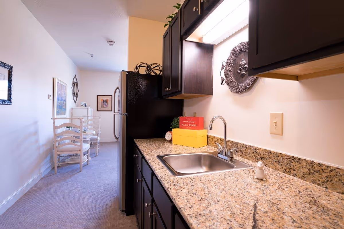 A small kitchen area with dark wooden cabinets, a granite countertop, and a stainless steel sink. There is a decorative plate hanging on the wall above the sink and a refrigerator at the end of the counter. In the background, there is a hallway with white chairs and framed artwork on the walls.
