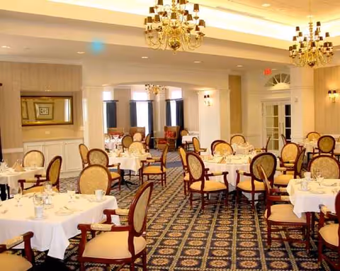 Elegant dining room with round tables, chairs, and chandeliers inside a senior living facility.