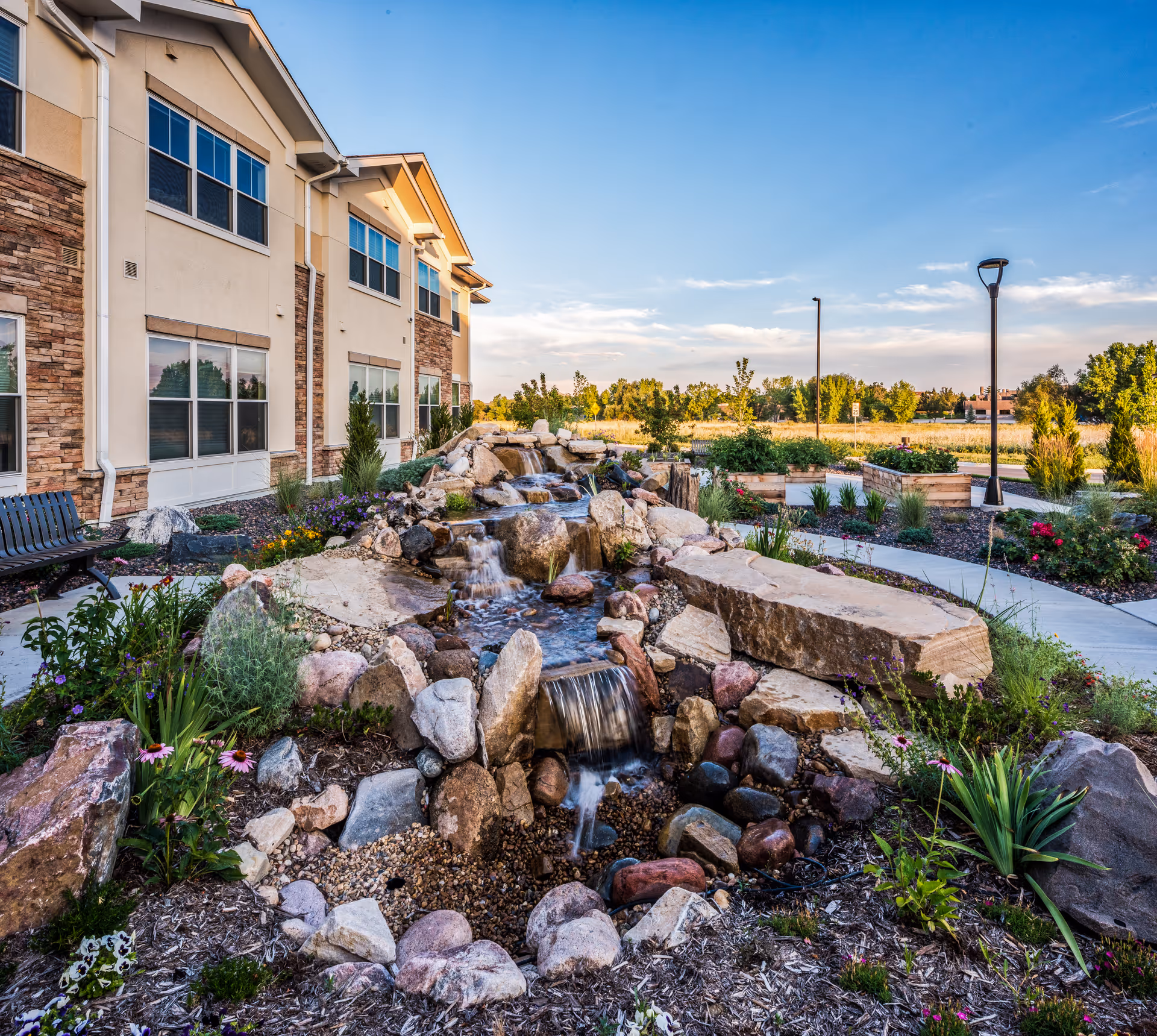Outdoor garden area at AltaVita Assisted Living featuring a small cascading rock waterfall surrounded by various plants and flowers, with a beige building on the left and a clear blue sky above.