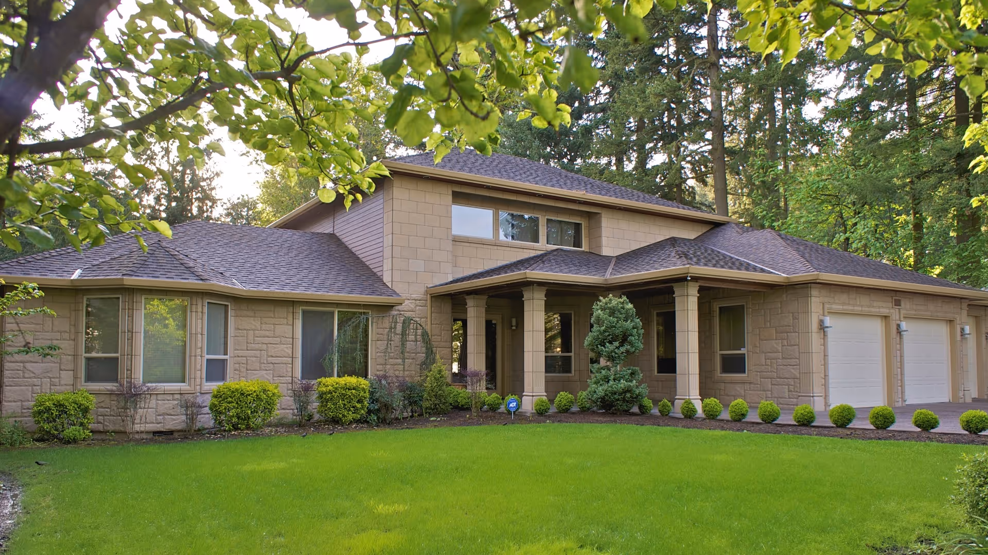 Exterior view of a single-story residential building with beige stone walls, a dark shingled roof, multiple windows, a three-car garage, and a well-maintained green lawn with shrubs and trees surrounding the property.