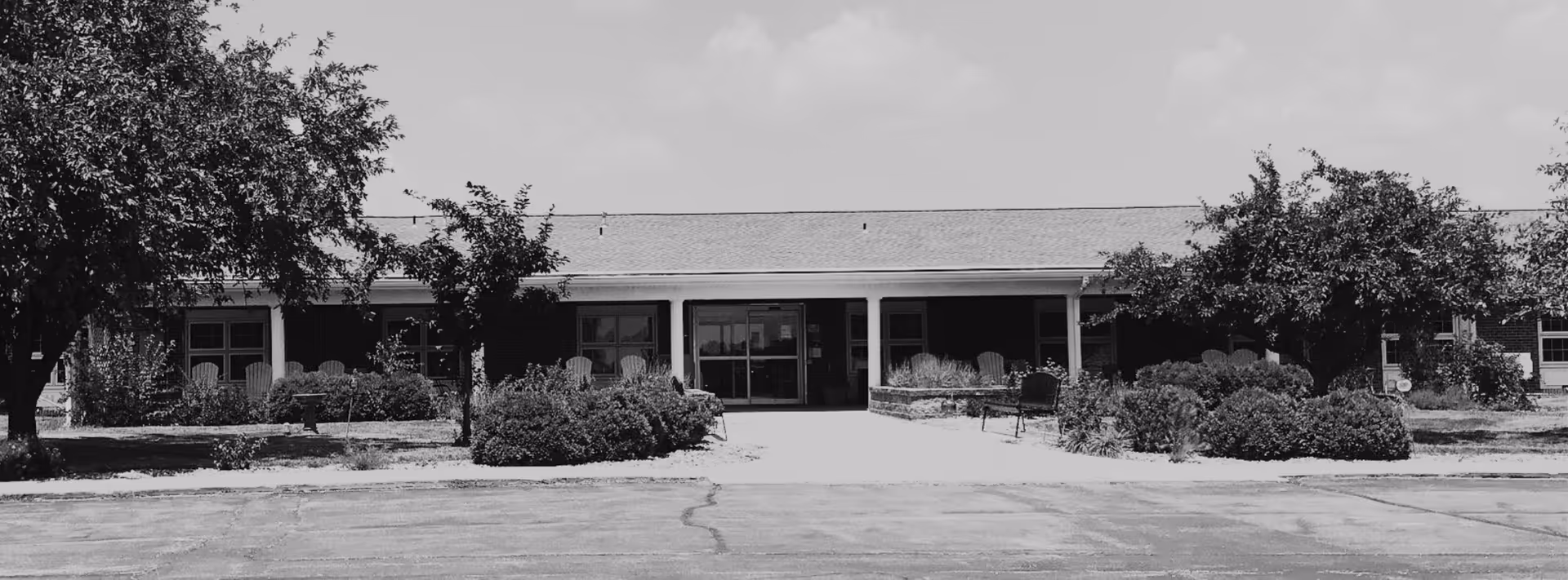 Black and white image showing the front exterior of a single-story building with a covered entrance, surrounded by trees and bushes, with a paved area in front.