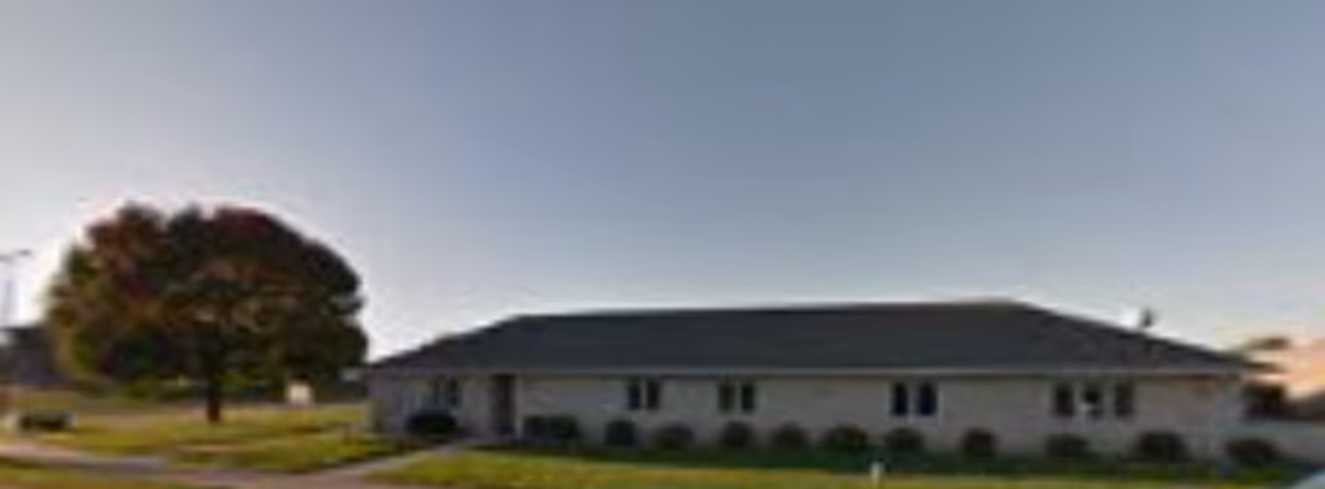 Single-story building with a dark roof and light-colored walls, surrounded by green grass and a large tree on the left side under a clear sky.