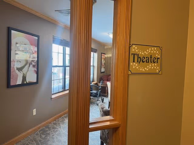 Interior view of a hallway leading to a room labeled 'Theater' with a decorative sign on the wall. The hallway has a framed artwork on the left wall and windows letting in natural light. Inside the theater room, there are chairs and a couch visible.