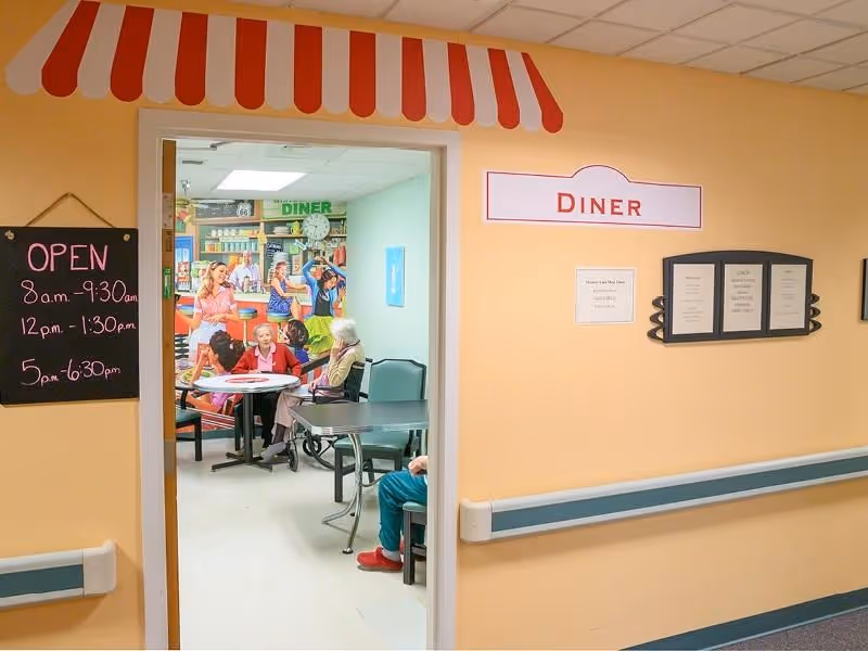 View through a doorway into a small diner area with tables and chairs. The walls are painted light yellow with a red and white striped awning decoration above the door. A chalkboard sign on the left lists diner open hours. Inside, elderly people are seated at tables, and a colorful mural depicting a classic diner scene is visible on the far wall. A sign above the door reads 'DINER'.