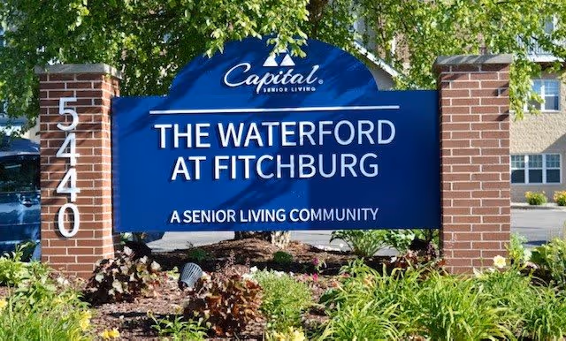 A blue sign with white text reading 'Capital Senior Living, The Waterford at Fitchburg, A Senior Living Community' mounted between two brick pillars with the number 5440 on the left pillar, surrounded by green plants and flowers with a building and trees in the background.