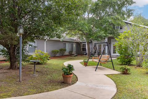 Outdoor garden area at Sumter Terrace Assisted Living featuring a curved concrete pathway, a wooden swing set, potted plants, a birdbath, and surrounding trees and shrubs with a gray building in the background under a partly cloudy sky.