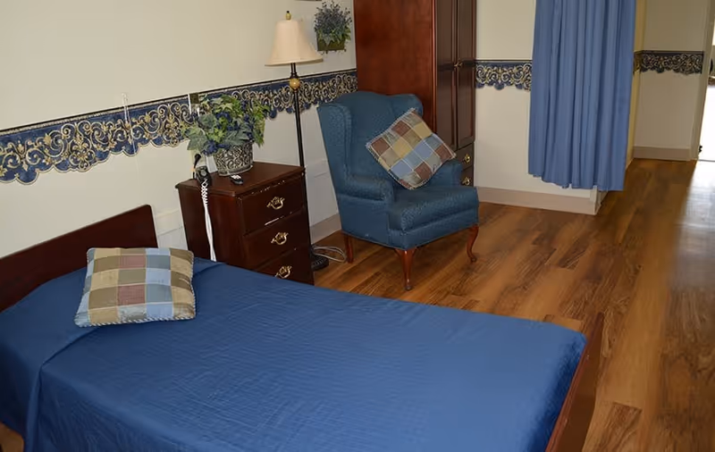 Single-occupancy bedroom with a blue-covered twin bed, wooden nightstand and lamp, upholstered armchair, and hardwood floor.