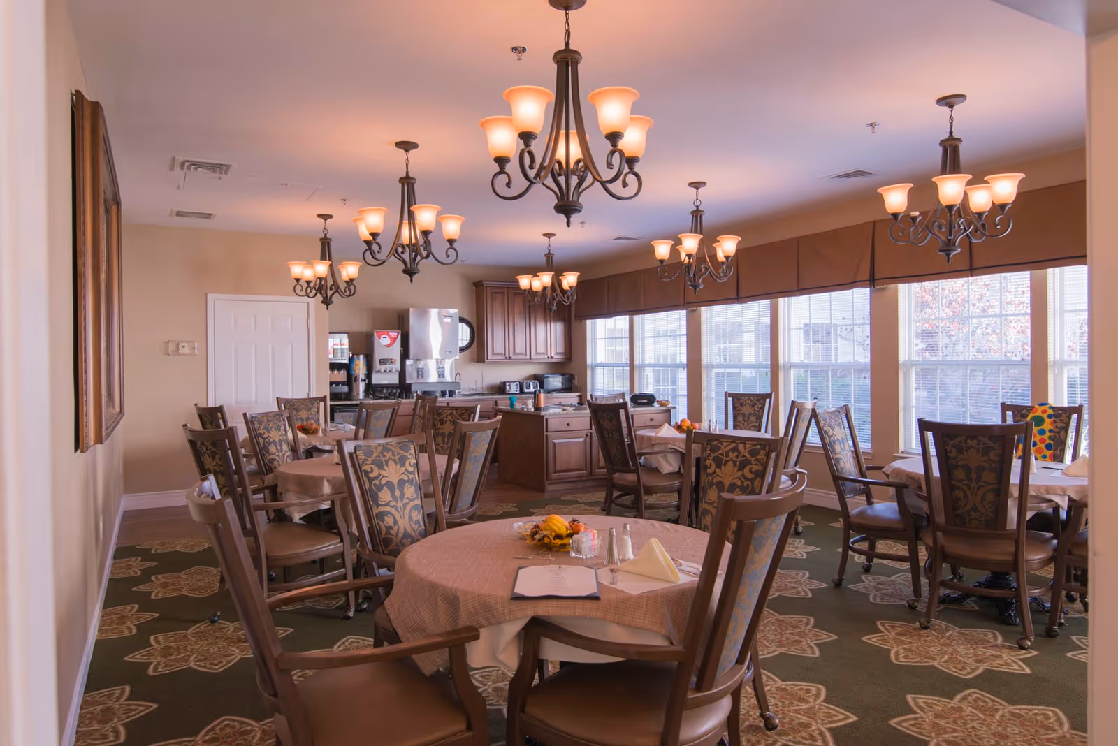Well-lit dining room with round tables set for meals, upholstered patterned chairs, multiple chandeliers, and large windows.