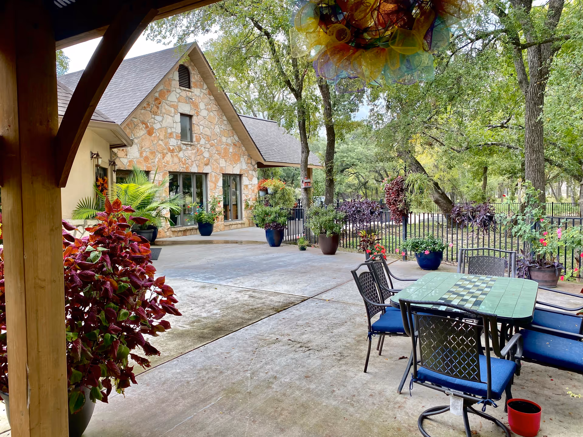Outdoor patio area at Wholehearted Care - New Hope Assisted Living featuring a stone building with large windows, surrounded by various potted plants and trees. There is a green table with a checkerboard pattern and four blue cushioned chairs on a concrete surface, with a black metal fence and more greenery in the background.