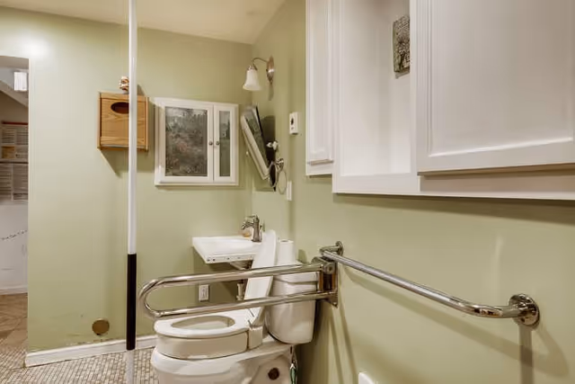 Accessible bathroom with a white toilet equipped with a safety grab bar on the right side, a small white sink with a faucet and a mirror above it, a wall-mounted light fixture, a wooden tissue holder, and a cabinet with white doors mounted on a light green wall.