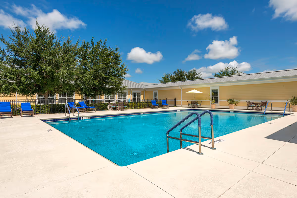 Outdoor swimming pool with metal handrails, blue lounge chairs, and a yellow single-story building under a sunny blue sky.