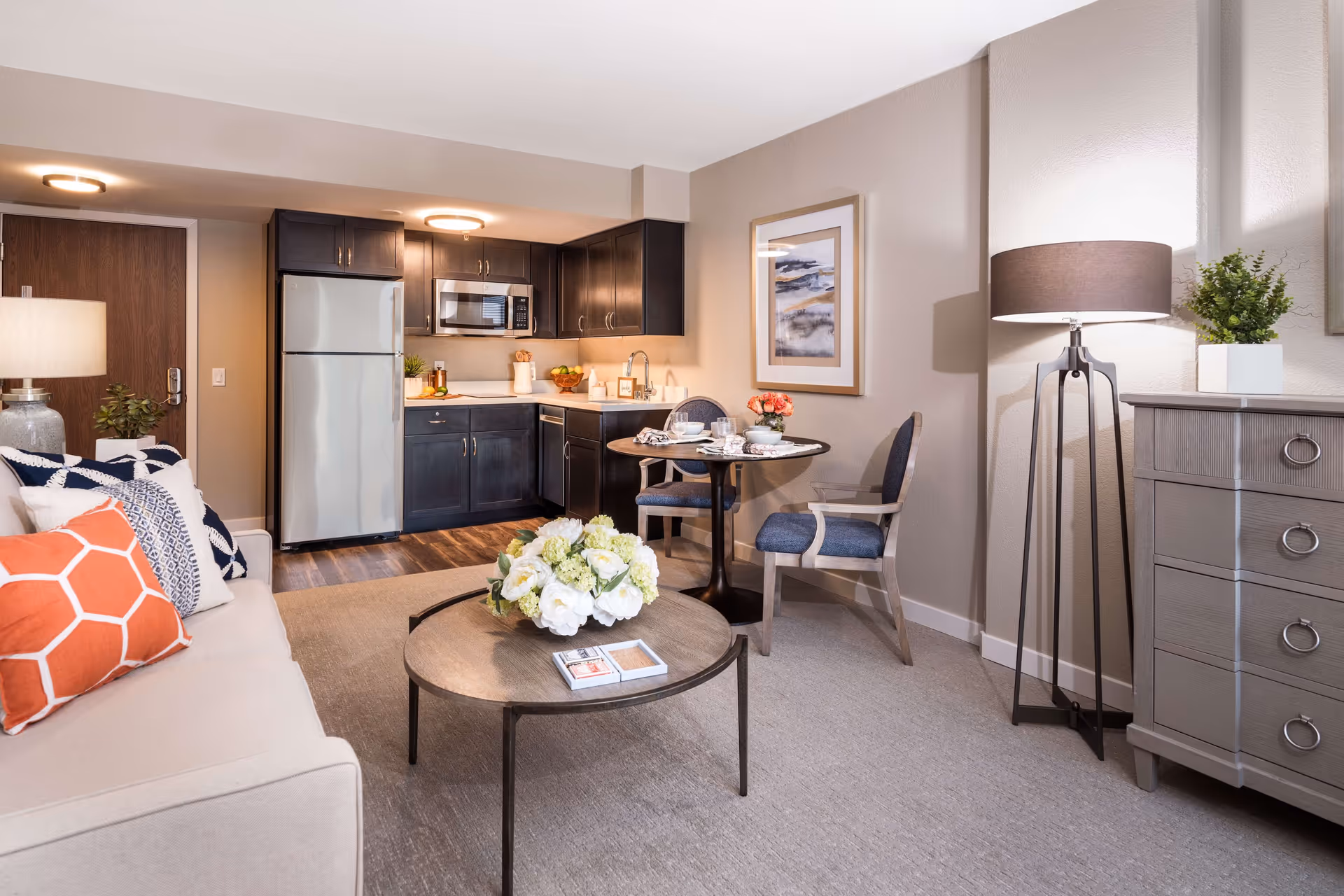 Well-lit studio living area with a sofa and coffee table, a small dining nook, and a kitchenette with dark cabinets and stainless appliances.