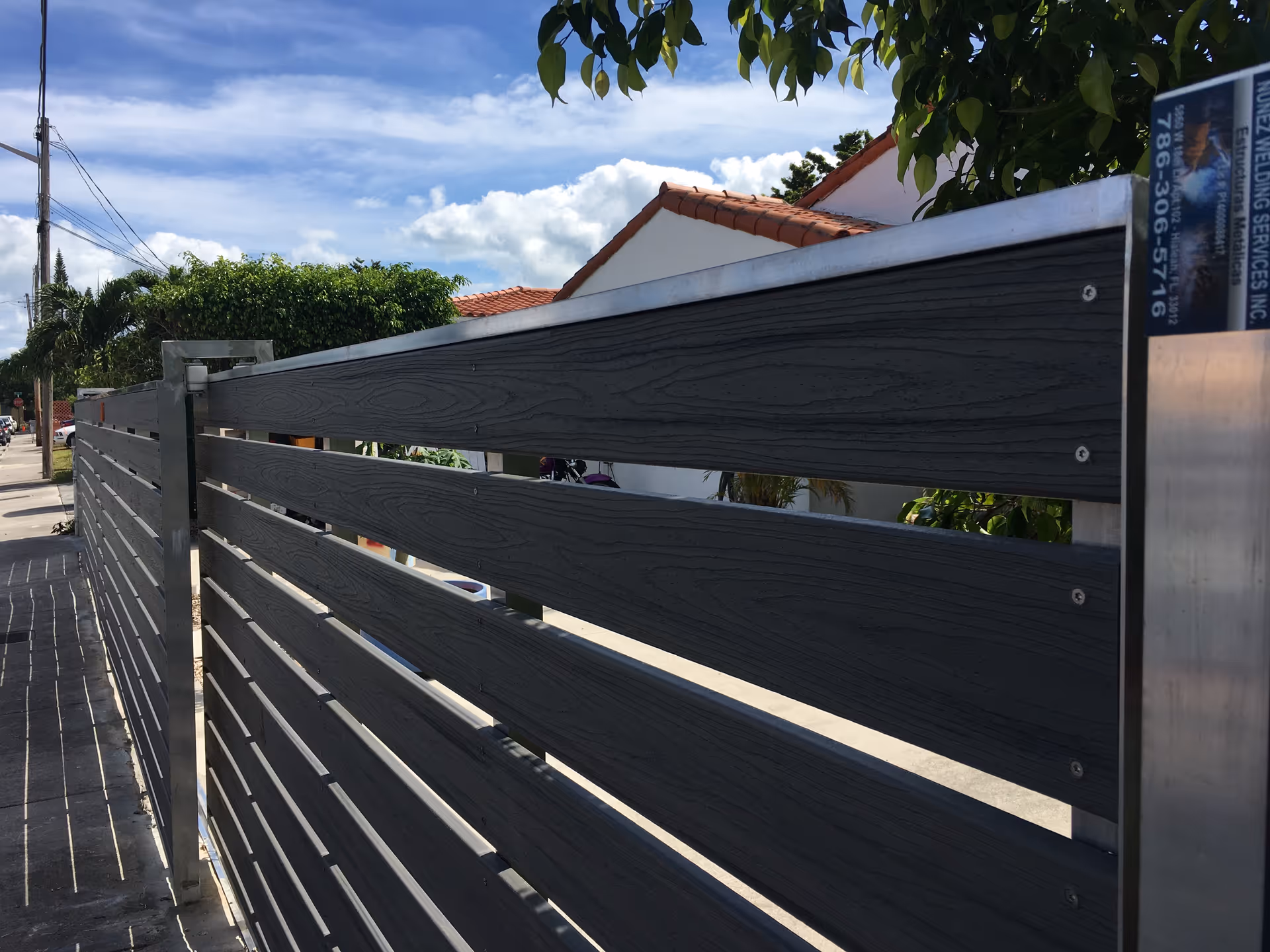 Modern horizontal slat metal fence and gate along a sidewalk with houses and trees visible behind it.