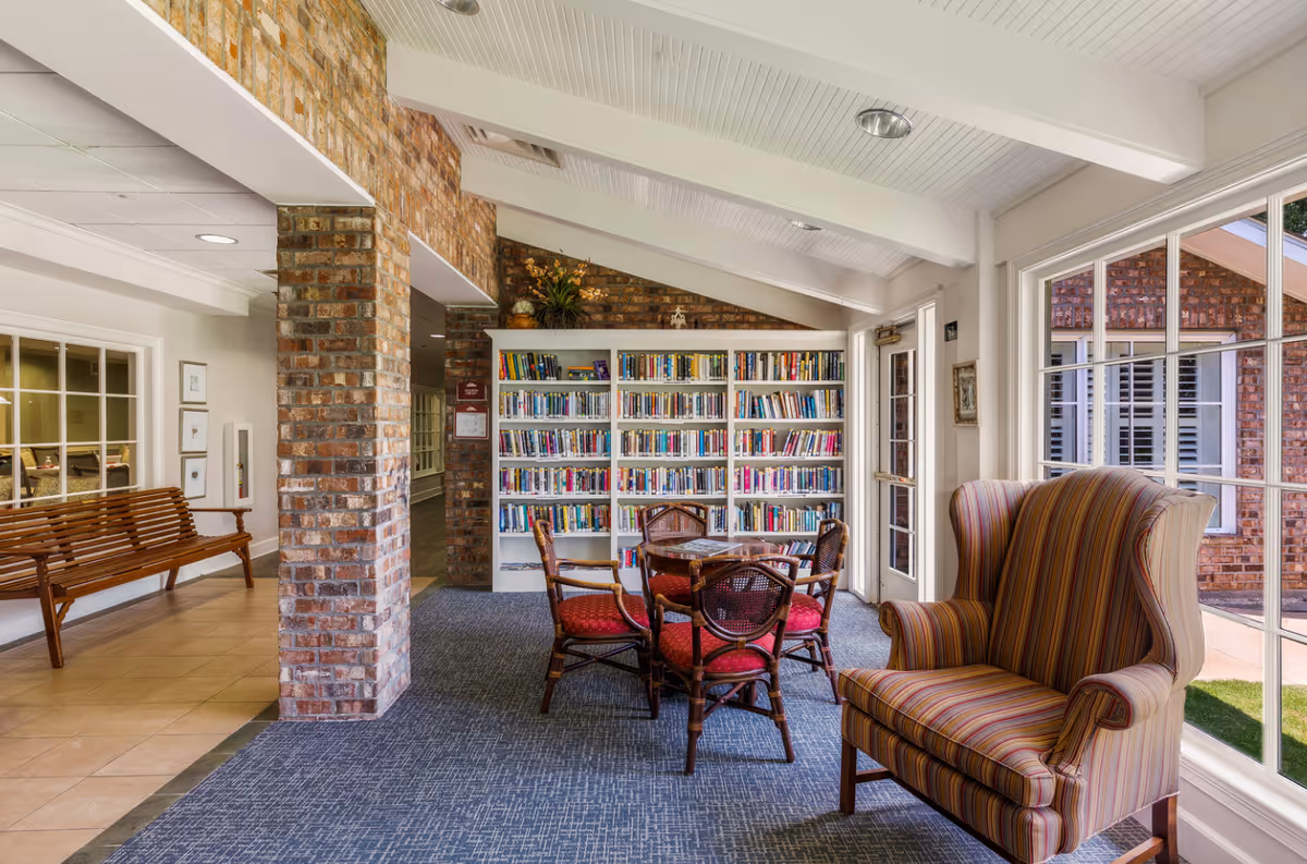 Sunlit common room with bookshelves, a table and chairs, and a striped armchair by large windows.