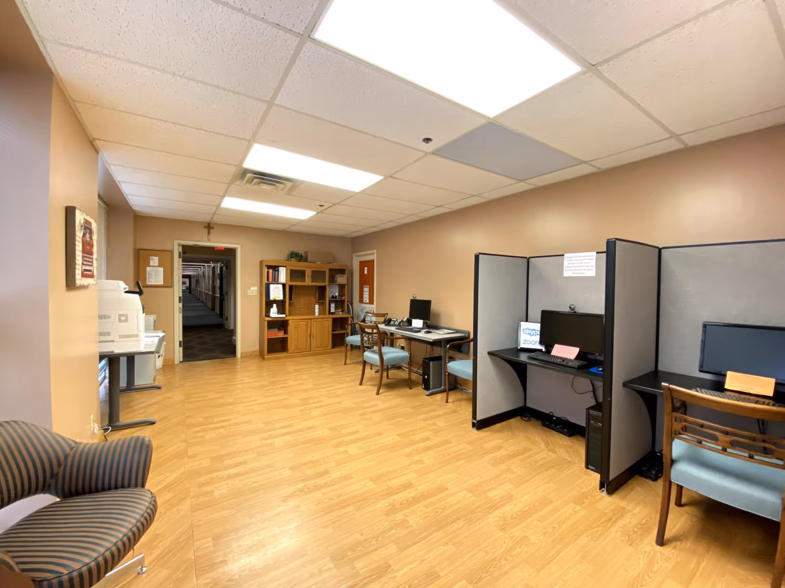 A room with several computer workstations separated by gray partitions, wooden chairs with blue cushions, a wooden cabinet with books and supplies, a printer on a small table, and a striped armchair. The room has beige walls, a light wood floor, and a drop ceiling with fluorescent lights.