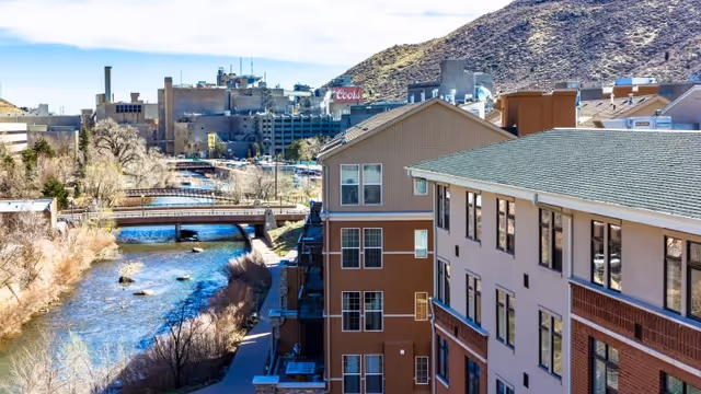 View of a river flowing alongside a multi-story building with a green roof and beige and brown exterior. In the background, there are industrial buildings and a mountain under a partly cloudy sky.