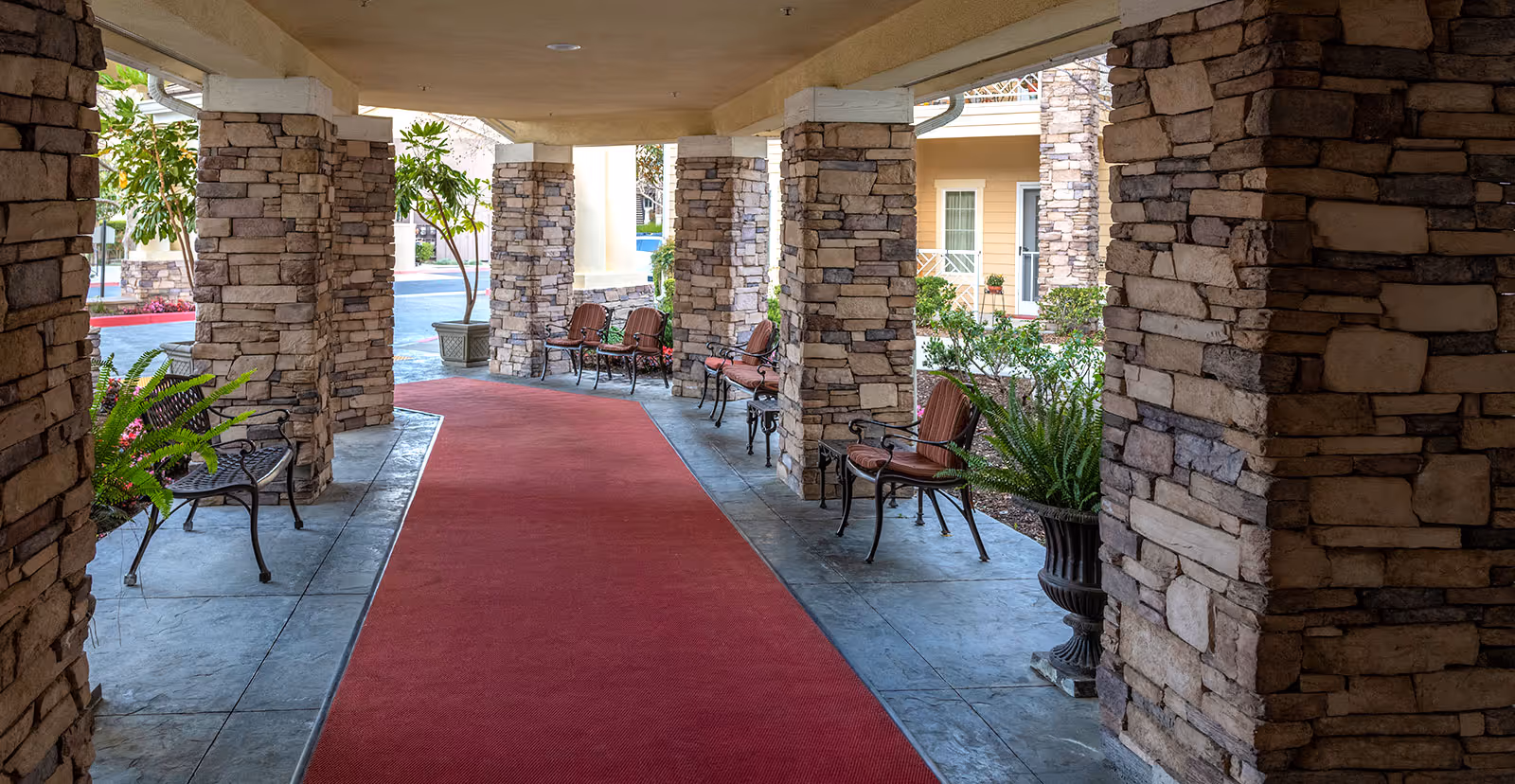 Covered outdoor walkway with stone pillars and a red carpet runner. Several benches with cushions and potted plants line the walkway. The background shows part of a building with windows and doors.