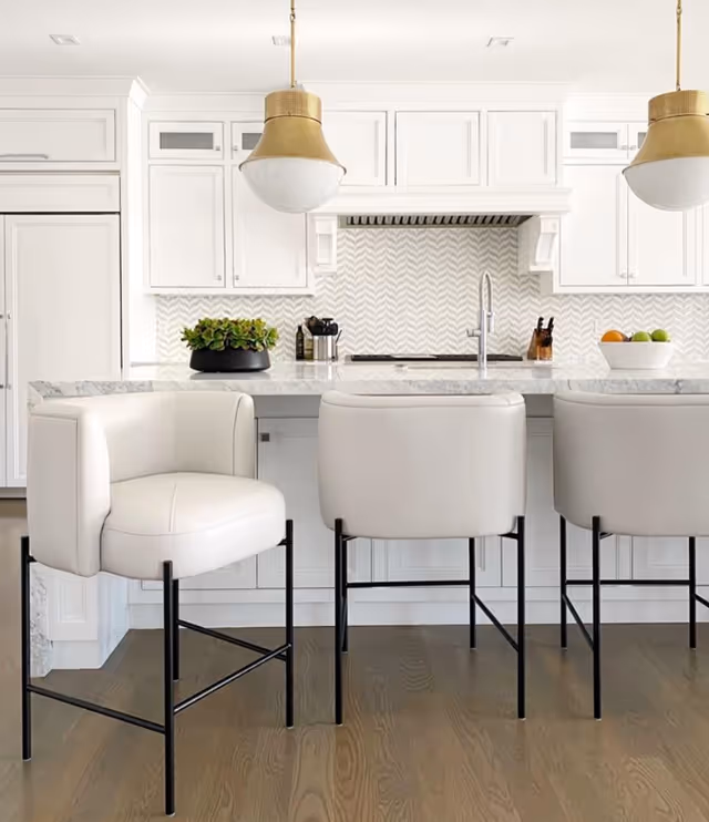 Bright white modern kitchen with a marble island, three white barstools, brass pendant lights, and a chevron tile backsplash.