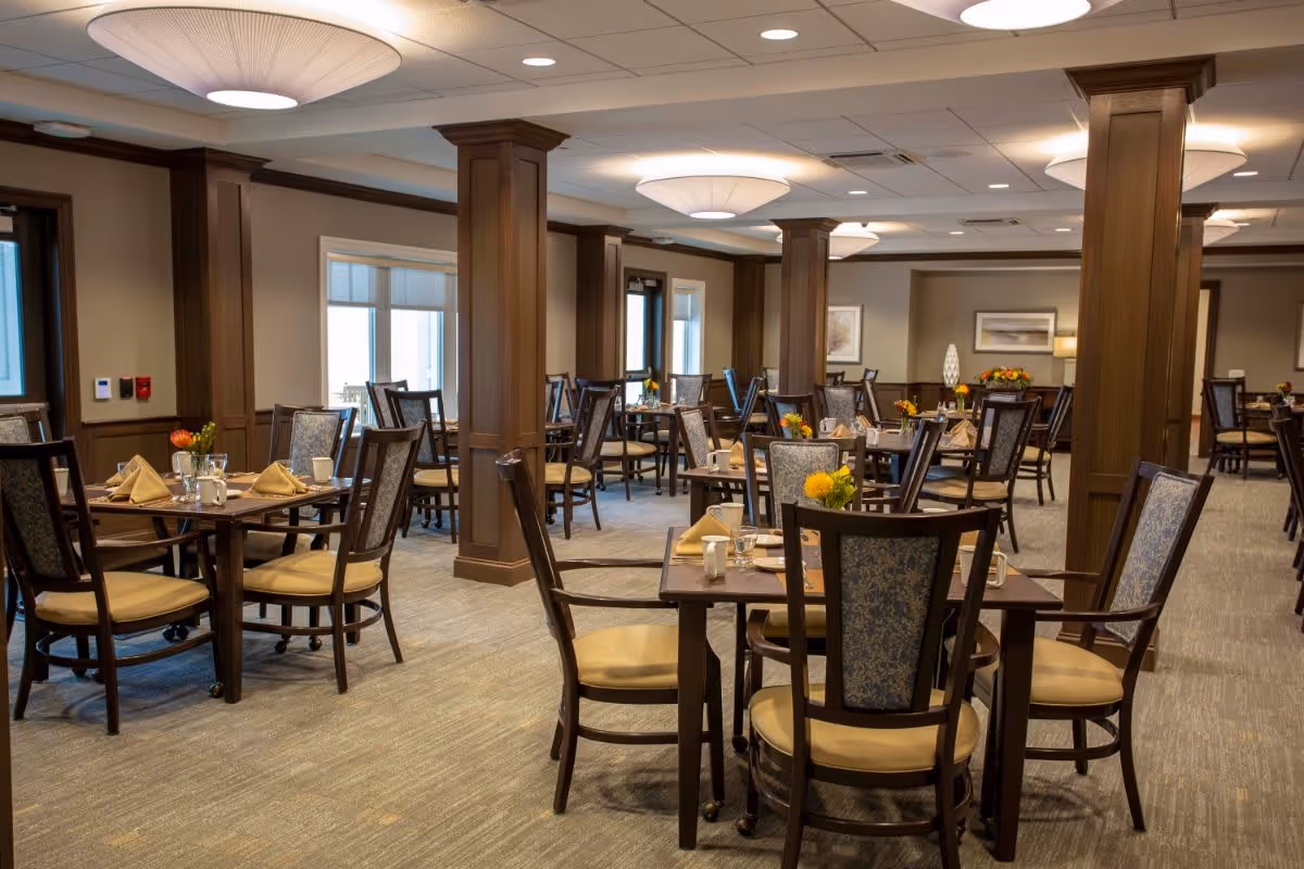 A spacious, well-lit dining room with multiple wooden tables and chairs arranged neatly. Each table is set with beige napkins, glassware, and small floral centerpieces. The room features wooden pillars, large windows with blinds, and soft overhead lighting fixtures.