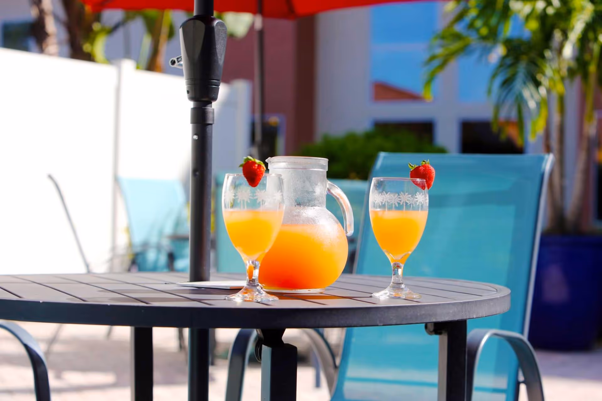 Outdoor patio table with a pitcher of orange juice and two glasses garnished with strawberries, surrounded by blue chairs and a red umbrella, with greenery and building in the background.
