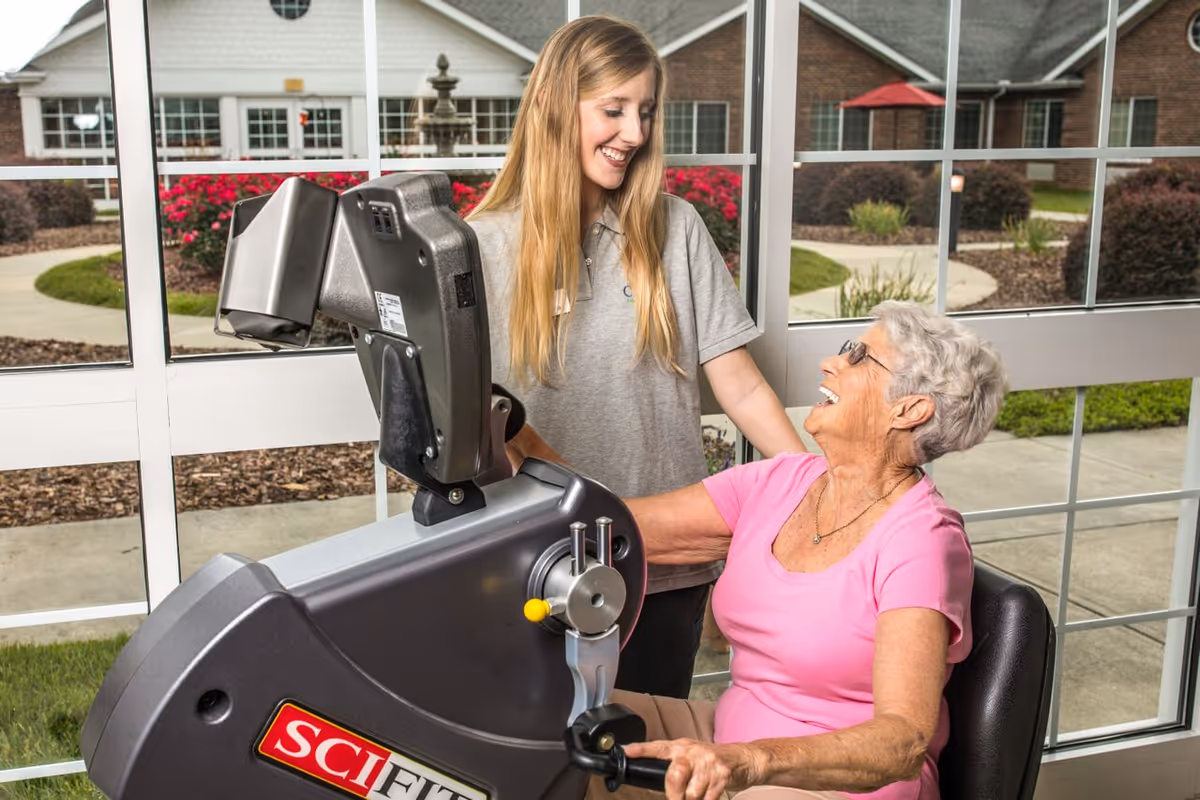 An elderly woman in a pink shirt is using a SCI FIT exercise machine indoors near large windows. A younger woman in a gray polo shirt is standing beside her, smiling and offering support. Outside the windows, there is a garden with bushes and a building in the background.