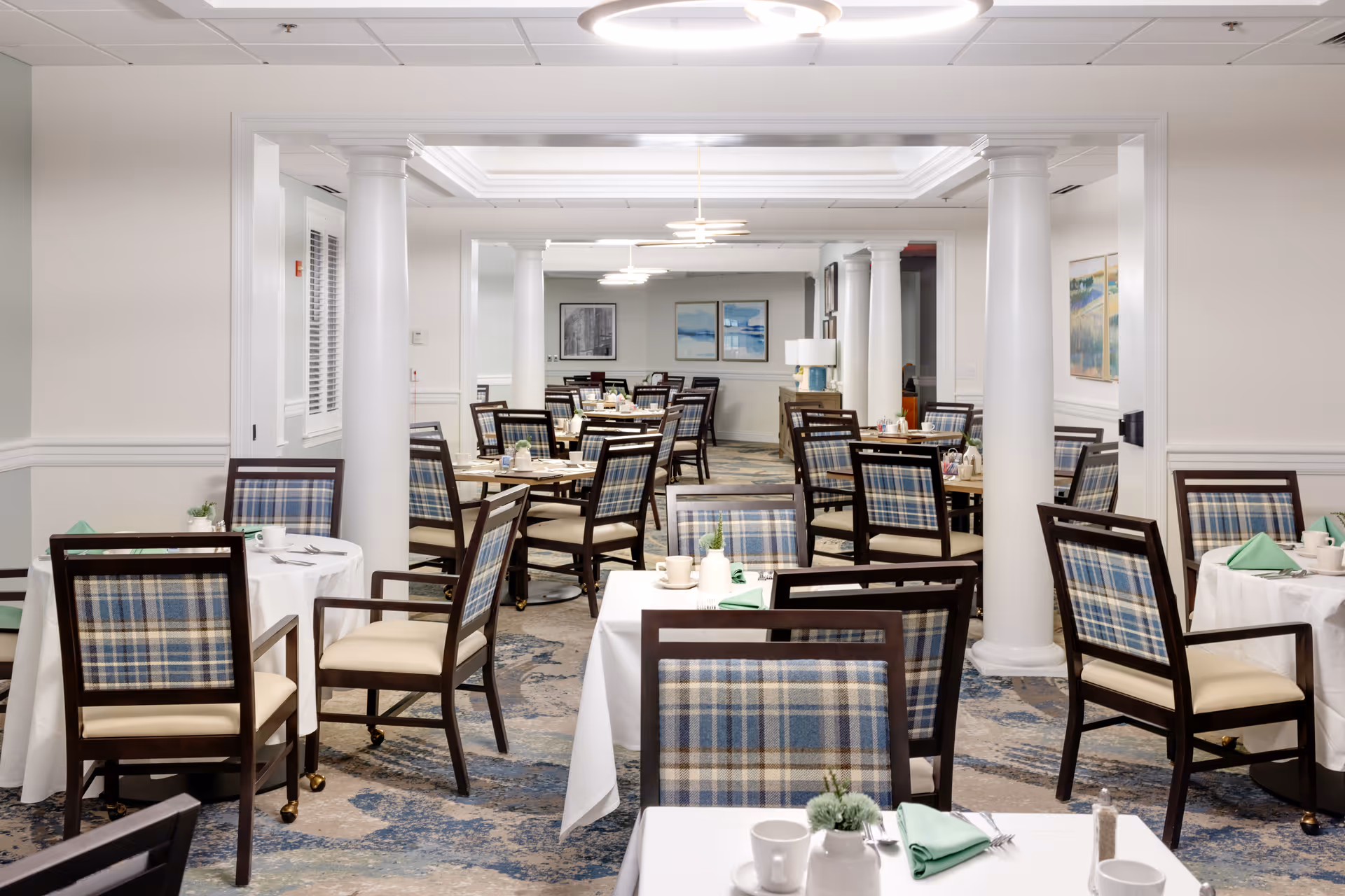 Bright, neatly arranged dining room with multiple tables and plaid-upholstered chairs in a senior living facility.