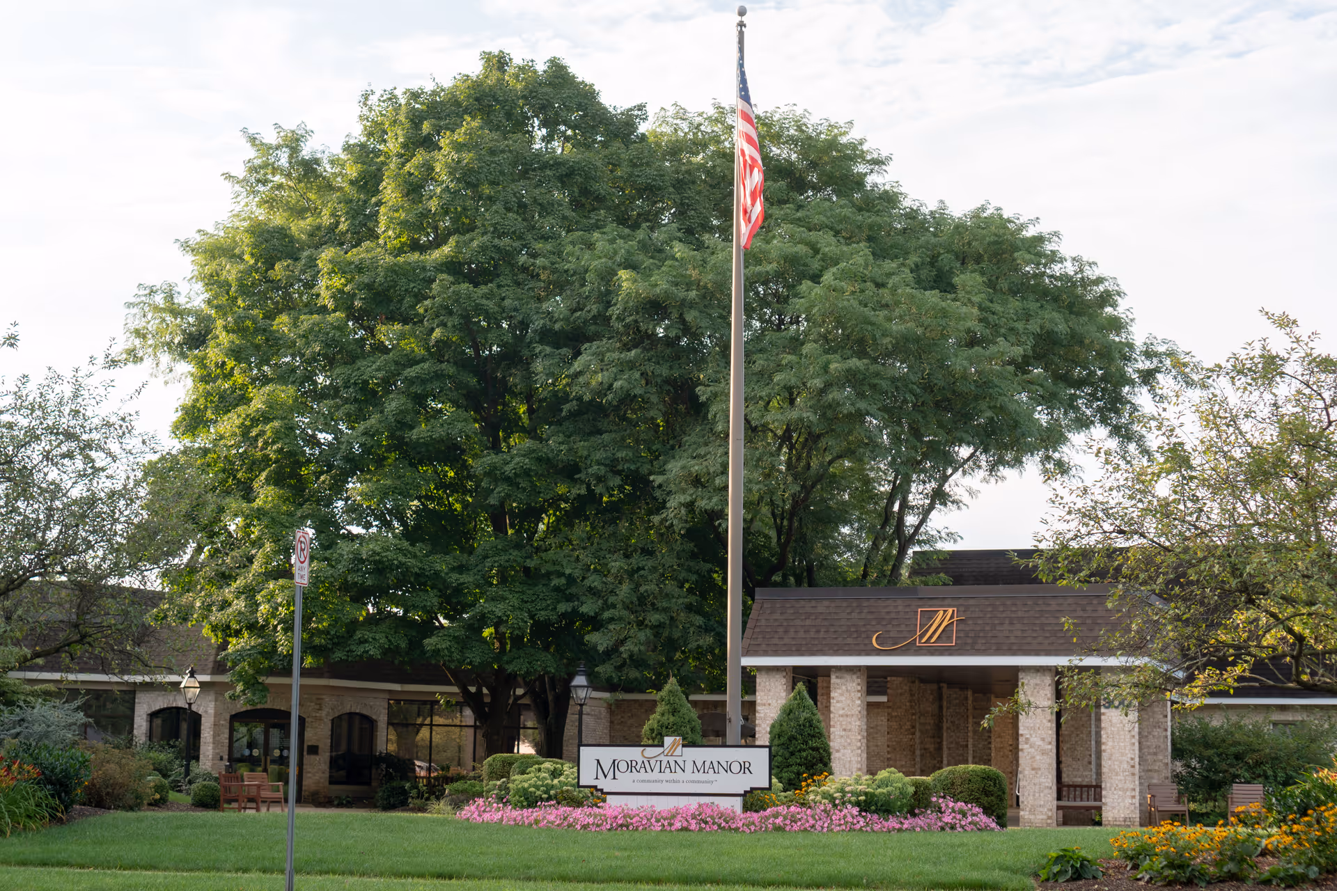 Exterior view of Moravian Manor Communities building with a large tree, an American flag on a flagpole, a sign reading 'Moravian Manor', and well-maintained landscaping with flowers and bushes.